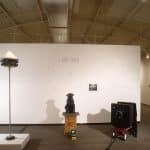Photo of front entrance exhibition space with Jno Cook name on wall. Three sculptures are in front. Left to right. Sculpture of tall pole with multiple cameras circling top of pole with white cone. Black dog sculpture with camera strapped around neck, sitting on wooden pedestal, with small video monitor on floor. Large black camera sitting on top of small red wagon.