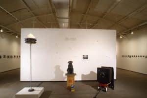 Photo of front entrance exhibition space with Jno Cook name on wall. Three sculptures are in front. Left to right. Sculpture of tall pole with multiple cameras circling top of pole with white cone. Black dog sculpture with camera strapped around neck, sitting on wooden pedestal, with small video monitor on floor. Large black camera sitting on top of small red wagon.