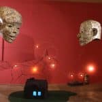 Photo of exhibition space, with mixed media sculptures. A unicorn shaped image made of lights and wire on back red wall. Two large head sculptures, one female and another male, hanging by ceiling, made of one dollar bills. Underneath is a small house, with a blue light illuminating inside, made from charcoal briquettes on top of material to resemble a lawn. At the bottom right against the wall is an old small decorative truck and on its right is a stack of small multi color dishes.