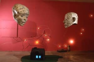 Photo of exhibition space, with mixed media sculptures. A unicorn shaped image made of lights and wire on back red wall. Two large head sculptures, one female and another male, hanging by ceiling, made of one dollar bills. Underneath is a small house, with a blue light illuminating inside, made from charcoal briquettes on top of material to resemble a lawn. At the bottom right against the wall is an old small decorative truck and on its right is a stack of small multi color dishes.