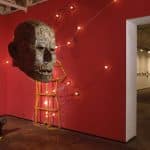 Photo of exhibition space, with mixed media sculptures. Large male head sculpture made of one dollar bills. Behind is a three-dimensional wooden wire frame of a human body from the neck to thighs and biceps on the back red wall. There is a sports car, a wolf, and a men’s formal suit shaped images made of lights and black wire on the back red wall surrounding the human wooden frame. The bottom left corner, on the floor, there is a glossy black hippopotamus head with a yellow light underneath peaked through the mouth, nostrils, and eyes.