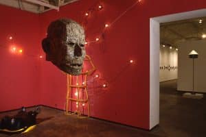 Photo of exhibition space, with mixed media sculptures. Large male head sculpture made of one dollar bills. Behind is a three-dimensional wooden wire frame of a human body from the neck to thighs and biceps on the back red wall. There is a sports car, a wolf, and a men’s formal suit shaped images made of lights and black wire on the back red wall surrounding the human wooden frame. The bottom left corner, on the floor, there is a glossy black hippopotamus head with a yellow light underneath peaked through the mouth, nostrils, and eyes.