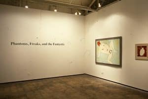 Corner of gallery, white walls with black text reading Phantoms, Freaks, and the Fantastic on the left. Right wall has two pieces of framed art. The first is a large painterly piece with a seagreen background, a white human figure laying down and leg spread, and the head is read. The next piece is small, with a red background, a white human figure with their back facing the viewer and scrunch downward.