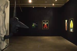 Inside a dark room, with dim lights on the pieces of painterly art on black walls. On the left is a large sculpture emerging from a mural piece of an unproportional human body doing a hand-stand. The head is creature-like with a large pointer nose, small beaded eyes, long face and neck, open mouth, and long pointed horn.