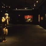 Inside a dark room, with dim lights on the pieces of painterly art on black walls. On the left of the image is a male human sculpture covering his face with his hands.