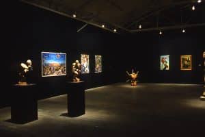Inside a dark room, with dim lights on the pieces of painterly art on black walls. There are three sculptures displayed across the room. The two on the left are on black pedestals. The middle sculpture is a human body resembling an uncomfortable squat. The farthest sculpture is a human body from the neck to shoulders and thighs, with six rods pocking out the body.