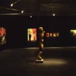 Inside a dark room, with dim lights on the pieces of art on black walls. The artwork on the wall are painterly, varying in sizes showing human like figures. In the center of the room, is an unrealistic proportionate human sculpture with dismembered arms.