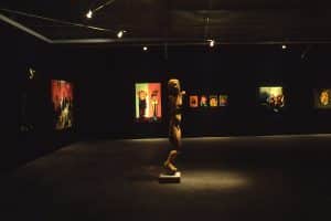 Inside a dark room, with dim lights on the pieces of art on black walls. The artwork on the wall are painterly, varying in sizes showing human like figures. In the center of the room, is an unrealistic proportionate human sculpture with dismembered arms.