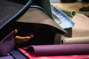 photograph of artist Preetika Rajgariah's artwork titled Waiting to Exhale. It is a close up photograph of numerous yoga mats of a variety of colors (blues, purples, reds, grays, and greens that are folded, rolled, and stacked into a mound with a ceramic hand reaching out from under them.