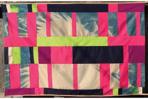 Artwork by Isela Aguirre: a cyanotype on cotton measuring 32 inches tall x 49 inches wide; the cyanotype of white feathers on blue background is pieced together like a quilt with cotton strips of vertical and horizontal rectangles of various sizes, forming an irregular grid. The cotton strips are various shades of white, pinks, bright green-yellow, magenta and black